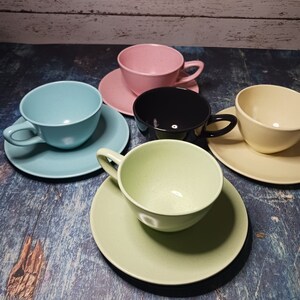 May include: Four teacups and saucers in different colors: light blue, light green, pink, and black. The cups and saucers are arranged on a blue wooden surface.