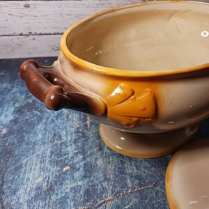 May include: A brown and beige ceramic serving bowl with a decorative handle and a yellow floral design. The bowl has a pedestal base and a matching lid.