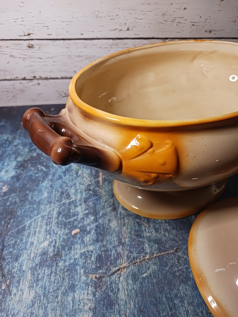May include: A brown and beige ceramic serving bowl with a decorative handle and a yellow floral design. The bowl has a pedestal base and a matching lid.