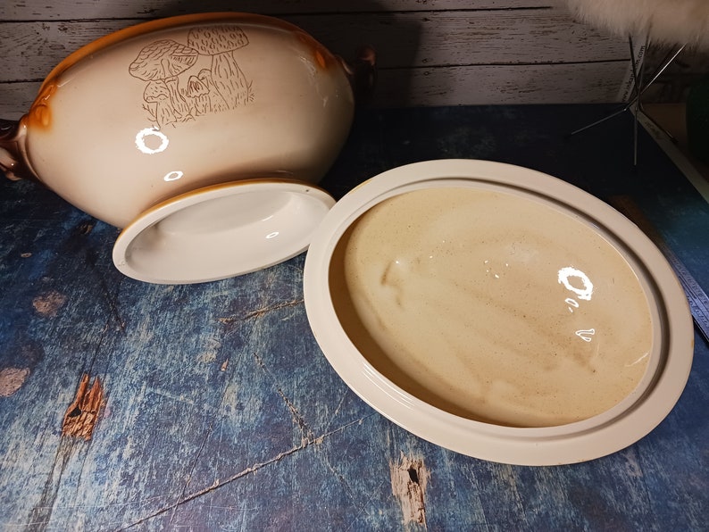 May include: A white ceramic serving bowl with a brown rim and a mushroom design. The bowl has a lid and a base.