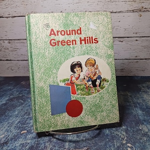 May include: Vintage children's book titled "Around Green Hills." The cover is light green with a dotted pattern and features an illustration of two children with a rabbit. The title is in red, and there is a blue square and red circle on the cover.