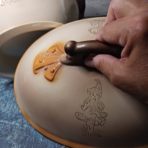 May include: A ceramic serving dish with a lid. The lid features a brown handle and a whimsical illustration of a gnome sitting on a mushroom. The dish has a similar illustration of mushrooms and trees.