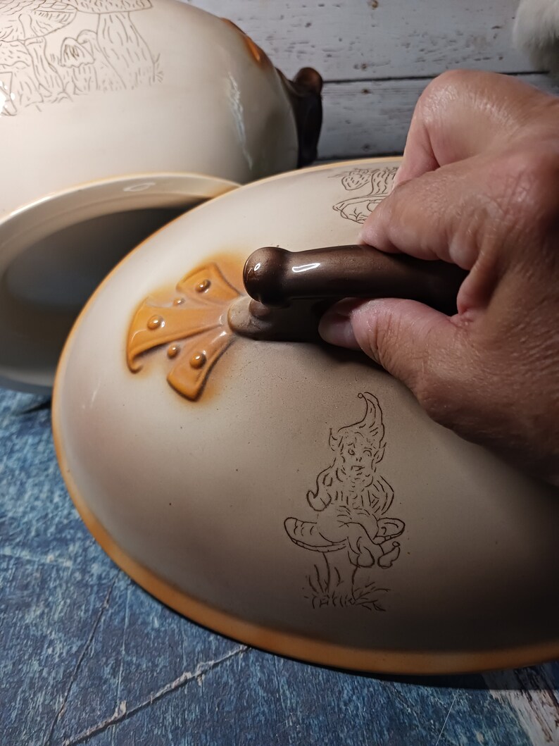 May include: A ceramic serving dish with a lid. The lid features a brown handle and a whimsical illustration of a gnome sitting on a mushroom. The dish has a similar illustration of mushrooms and trees.
