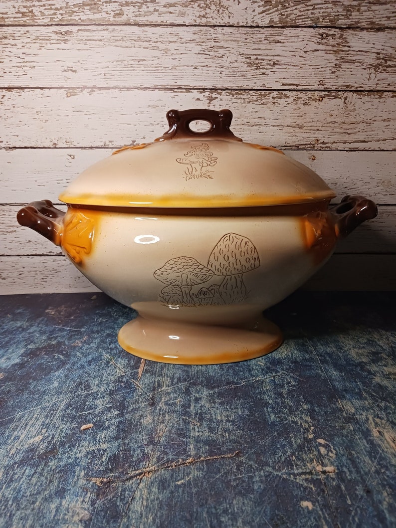 May include: A ceramic serving bowl with a lid, featuring a mushroom design in brown and white on a beige background. The bowl has two brown handles and a pedestal base.
