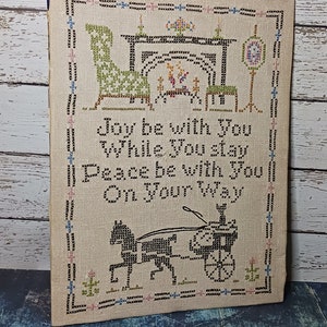 May include: A framed cross-stitch sampler with the text "Joy be with You While You Stay Peace be with You On Your Way." The artwork features a horse-drawn carriage, furniture, and floral accents, all in a neutral color palette.