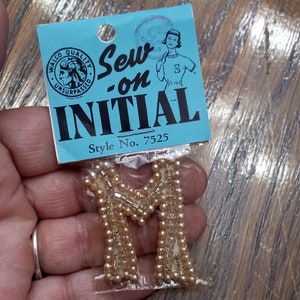 May include: A beige fabric letter M, decorated with small white beads, is packaged in a clear plastic bag with a blue and white label that reads "Sew-on INITIAL Style No. 7525".