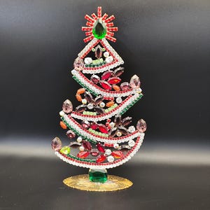 May include: A decorative Christmas tree figurine. The tree is adorned with red, green, and clear gemstones, with a large green gem at the top. The base is gold-colored, and the tree is set against a black background.