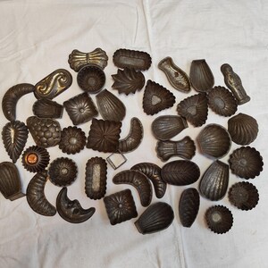 Op de afbeelding: Een verzameling vintage metalen koekjessnijders in diverse vormen, waaronder bloemen, schelpen en geometrische ontwerpen. De snijders zijn gemaakt van donker, verouderd metaal en liggen op een witte ondergrond. De collectie bevat verschillende maten en stijlen.