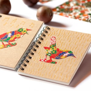 Peut inclure: Deux cahiers à spirales avec des couvertures en papier kraft. Chaque cahier présente un motif d'oiseau coloré avec des motifs floraux rouges, jaunes, bleus et verts. Des notes de musique sont imprimées au-dessus d'un oiseau. Les cahiers sont sur une surface blanche.