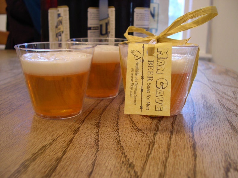 May include: Clear plastic cups filled with amber-colored soap, resembling beer, with a white foamy top. One soap is wrapped with a yellow ribbon and a tag that reads "MAN CAVE BEER Soap for Men". The soap is on a wooden surface.