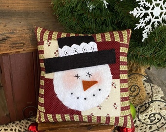 May include: A handmade snowman cushion with a red and white striped background. The cushion is sitting on a wooden sledge with a red bauble.
