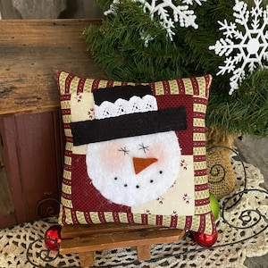Puede incluir: Una almohada de muñeco de nieve hecha a mano con un fondo a rayas rojo y blanco. La almohada está sentada en un trineo de madera con un adorno rojo.