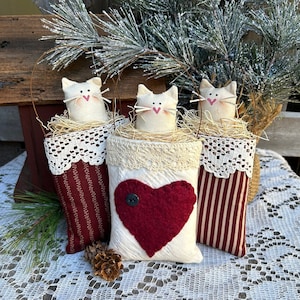 Pode incluir: Três ornamentos de gato de feltro branco com corações vermelhos, aninhados em sacos de juta e tecido com detalhes em renda. Os sacos são pendurados com laços de arame e têm um estilo rústico e de fazenda.