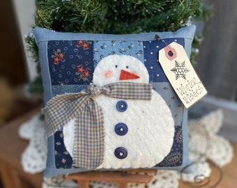May include: A blue and white patchwork pillow with a snowman design. The snowman has a red nose, black eyes, and three black buttons. The pillow has a blue and white plaid ribbon around its neck and a tag that says "Winter Wishes".