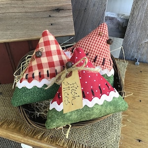 May include: Three handmade watermelon-shaped pillows in a basket. The pillows are made of red and white gingham fabric, red fabric with white polka dots, and green fabric. The pillows have white stitching around the edges and black stitching to resemble watermelon seeds. A small brown tag with the text "Fresh Slices" is attached to one of the pillows.
