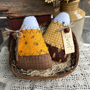 May include: Two handmade corn-shaped fabric decorations in a woven basket. One features a yellow and brown acorn pattern, the other a yellow gingham pattern. Both have white tops and are stitched with brown thread. One has a tag that says "Harvest".