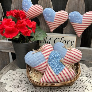 May include: A collection of patriotic heart-shaped ornaments with red and white striped fabric and blue star-patterned fabric. Some hearts are hanging on a wooden fence, while others are in a wicker basket. A sign reads "Old Glory".