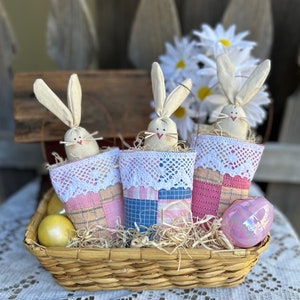 May include: Three handmade bunny-shaped Easter baskets in a wicker basket. The baskets are made of fabric with a patchwork design and lace trim. The baskets are filled with straw and decorated with pastel-colored Easter eggs.