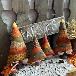 Op de afbeelding: Vier kegelvormige stoffen bomen in herfstkleuren zoals oranje, bruin en groen met diverse blad- en bloemmotieven. Elke boom staat op een kleine houten voet, waarbij twee bomen zijn voorzien van een beige jute strik en een donkere knoop. Een houten bordje toont de tekst HARVEST.