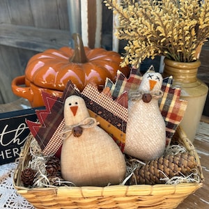 May include: Two handmade fabric turkeys with patchwork wings in a woven basket filled with pine cones and straw. The turkeys are white with brown accents and have black eyes and a red beak.