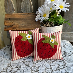 Może przedstawiać: Dwie czerwono-zielone poduszki z filcu w kształcie truskawek z białymi kwiatowymi akcentami na czerwono-białym paskowanym materiale. Poduszki leżą na białym koronkowej serwetce.