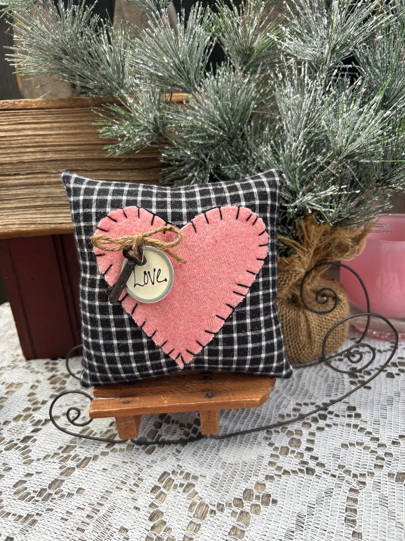 Peut inclure: Un petit coussin en tissu &agrave; carreaux noirs et blancs avec un c&oelig;ur en feutre rose cousu sur le devant. Le c&oelig;ur est orn&eacute; d'une cl&eacute; et d'une &eacute;tiquette sur laquelle est inscrit "Love". Le coussin est pos&eacute; sur un petit tra&icirc;neau en bois.