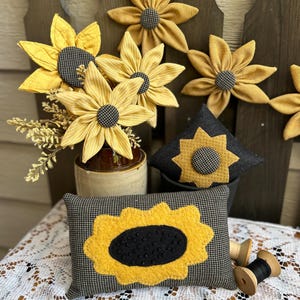 May include: A collection of fabric sunflower decorations including a rectangular pillow with a black and yellow sunflower applique, a diamond-shaped pillow, and several fabric sunflowers on stems in a container, featuring yellow and black patterns.