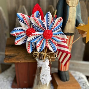 May include: Patriotic fabric flower bouquet in a clear glass vase. The flowers feature red, white, and blue striped and starred petals with red polka dot centers. The vase is adorned with lace and tied with twine. A wooden Uncle Sam figure is in the background.