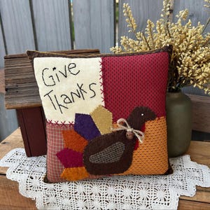 May include: A square Thanksgiving pillow with a patchwork design. The pillow features a turkey applique and the words "Give Thanks" embroidered on a cream-colored patch. The turkey has a brown body, colorful tail feathers, and a small bow.