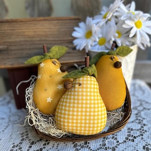 May include: Three stuffed fabric pears in a small woven basket. One pear features a yellow star pattern, one has a yellow and white gingham check, and one is solid mustard yellow. Each pear has a brown stem, green felt leaves, and a small bell.
