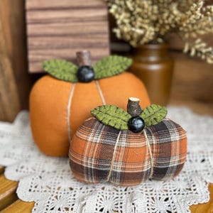 May include: Two fabric pumpkins, one solid orange and one orange, black, and white plaid. Each features green felt leaves, a dark button at the center, and a brown stem. The pumpkins are round and segmented with visible stitching lines.