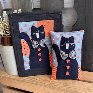 May include: Two fabric items featuring black cat appliqués. The larger rectangular piece has a black border and a light blue background with orange pumpkins. The smaller rectangular pillow has a light blue pumpkin background and orange striped sides.