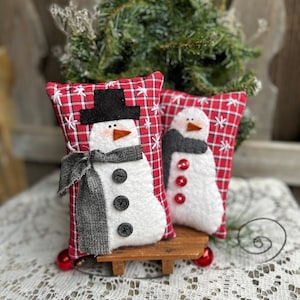 May include: Two handmade snowman pillows with red and white plaid fabric, black buttons, and black hats. The snowmen are sitting on a wooden sled.