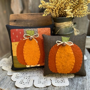 May include: Two handmade felt pumpkin pillows, one with a patchwork design and the other with a black and white plaid design. Both pillows feature a brown felt stem and a brown button.