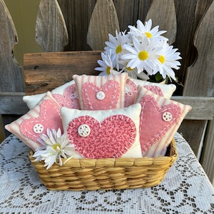 May include: A wicker basket filled with handmade felt heart-shaped cushions. The cushions are in shades of pink and white with button accents. The basket is sitting on a white lace tablecloth.