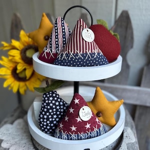 May include: A two-tiered white tray holds patriotic-themed fabric decorations. The top tier features cone-shaped items with red, white, and blue patterns, a red pear, and a yellow star. The bottom tier has a pear-shaped item with white stars and a cone with a "Liberty" tag.