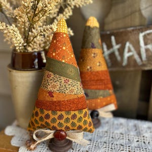 May include: Two cone-shaped fabric ornaments featuring horizontal patchwork stripes in shades of orange, olive green, and yellow with floral and leaf patterns. Each ornament sits on a small brown wooden base with a burlap bow and a button.