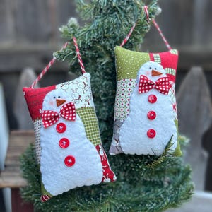 May include: Two handmade Christmas ornaments shaped like snowmen. Each snowman has a white felt body, a red and white checkered bow tie, and red button accents. The ornaments are hanging from red and white twine.