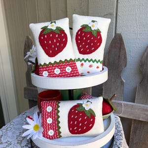 May include: Three decorative strawberry-themed pillows on a tiered tray. The pillows are white with a large red strawberry applique and green leaves. One pillow has a red and white polka dot border with white buttons. A white daisy is on the tray.