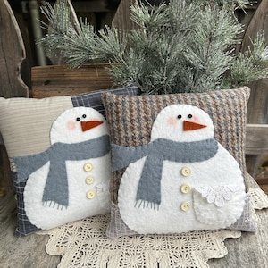 Puede incluir: Dos cojines decorativos con forma de muñeco de nieve. Cada cojín presenta un muñeco de nieve blanco con bufanda gris, nariz naranja y detalles de botones. Los cojines descansan sobre una superficie de madera con un mantel de encaje.