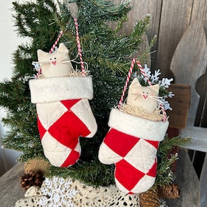 May include: Two white cat ornaments hanging from a Christmas tree. The ornaments are made of fabric and are shaped like mittens. The mittens are red and white with a diamond pattern. The cats are peeking out of the top of the mittens.