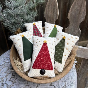 May include: A wooden bowl holds six Christmas tree-shaped pillows. The pillows are white with embroidered star accents and feature green, red, and plaid tree appliqués. Each tree is topped with a yellow star and a black button at the base.