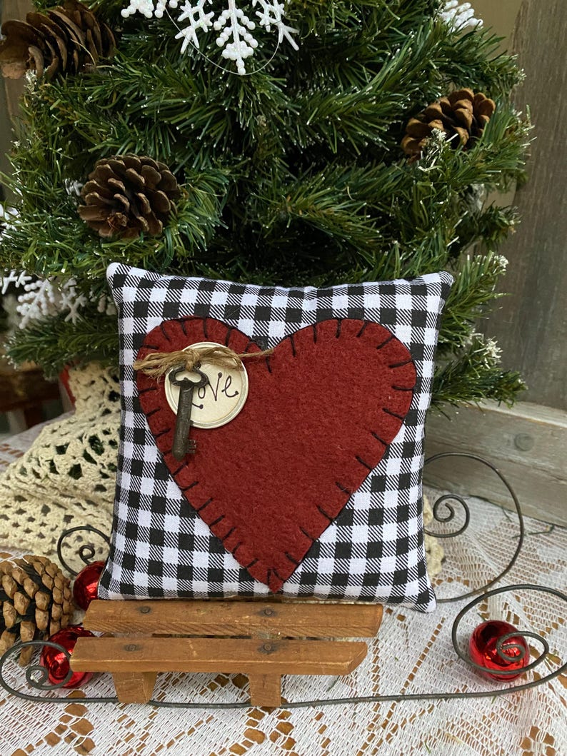 Peut inclure: Un coussin en forme de c&oelig;ur en feutre rouge avec une cl&eacute; et le mot "Love" sur un coussin &agrave; carreaux noirs et blancs. Le coussin est pos&eacute; sur un tra&icirc;neau en bois.