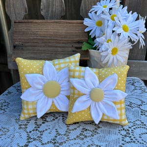 May include: Two small yellow and white gingham and polka dot pillows with white daisy appliques. The pillows are sitting on a white lace tablecloth.