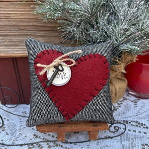 Peut inclure: Un petit coussin en forme de c&oelig;ur gris et rouge avec une cl&eacute; et le mot &laquo;&nbsp;LOVE&nbsp;&raquo; sur une &eacute;tiquette blanche. Le coussin est pos&eacute; sur un petit tra&icirc;neau en bois.