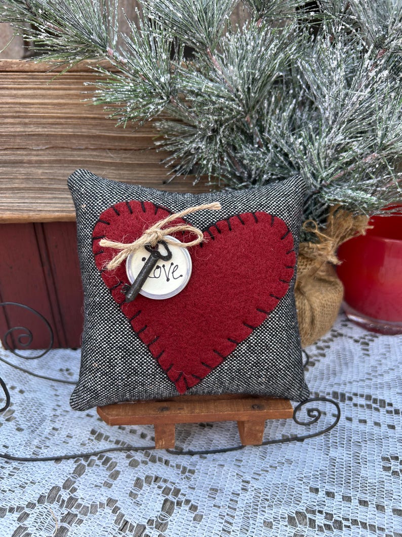 Peut inclure: Un petit coussin en forme de c&oelig;ur gris et rouge avec une cl&eacute; et le mot &laquo;&nbsp;LOVE&nbsp;&raquo; sur une &eacute;tiquette blanche. Le coussin est pos&eacute; sur un petit tra&icirc;neau en bois.