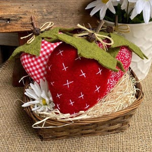 May include: A woven basket holds three heart-shaped strawberry decorations. The strawberries are red with white cross-stitch details and green felt leaves. One strawberry has a red and white gingham pattern. A white daisy and straw fill the basket.