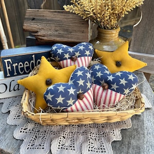 May include: A woven basket holds patriotic-themed decorative items. There are yellow felt stars and heart-shaped pillows with blue star-patterned fabric and red and white striped fabric. The word "Freedom" is visible on a blue wooden sign.