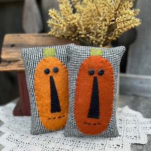 Peut inclure: Deux citrouilles en tissu à carreaux noirs et blancs avec des visages en feutre orange et des nez en feutre noir. Les citrouilles sont posées sur une dentelle blanche au crochet.
