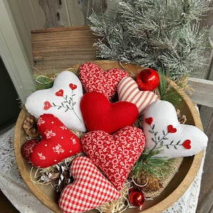 Puede incluir: Un cuenco de madera lleno de diversas decoraciones de tela en forma de corazón. Los corazones tienen patrones rojos y blancos, algunos con bordados florales. Piñas y vegetación se suman al tema rústico del Día de San Valentín. Un adorno rojo y una ramita de pino helado completan el arreglo.